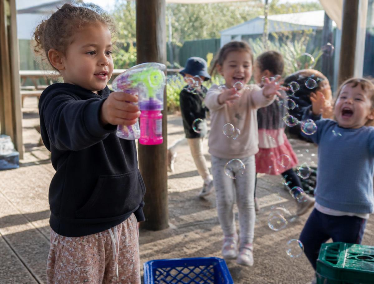 A child holding a bubble gun outdoors while other children run and reach for the bubbles in the morning sun
