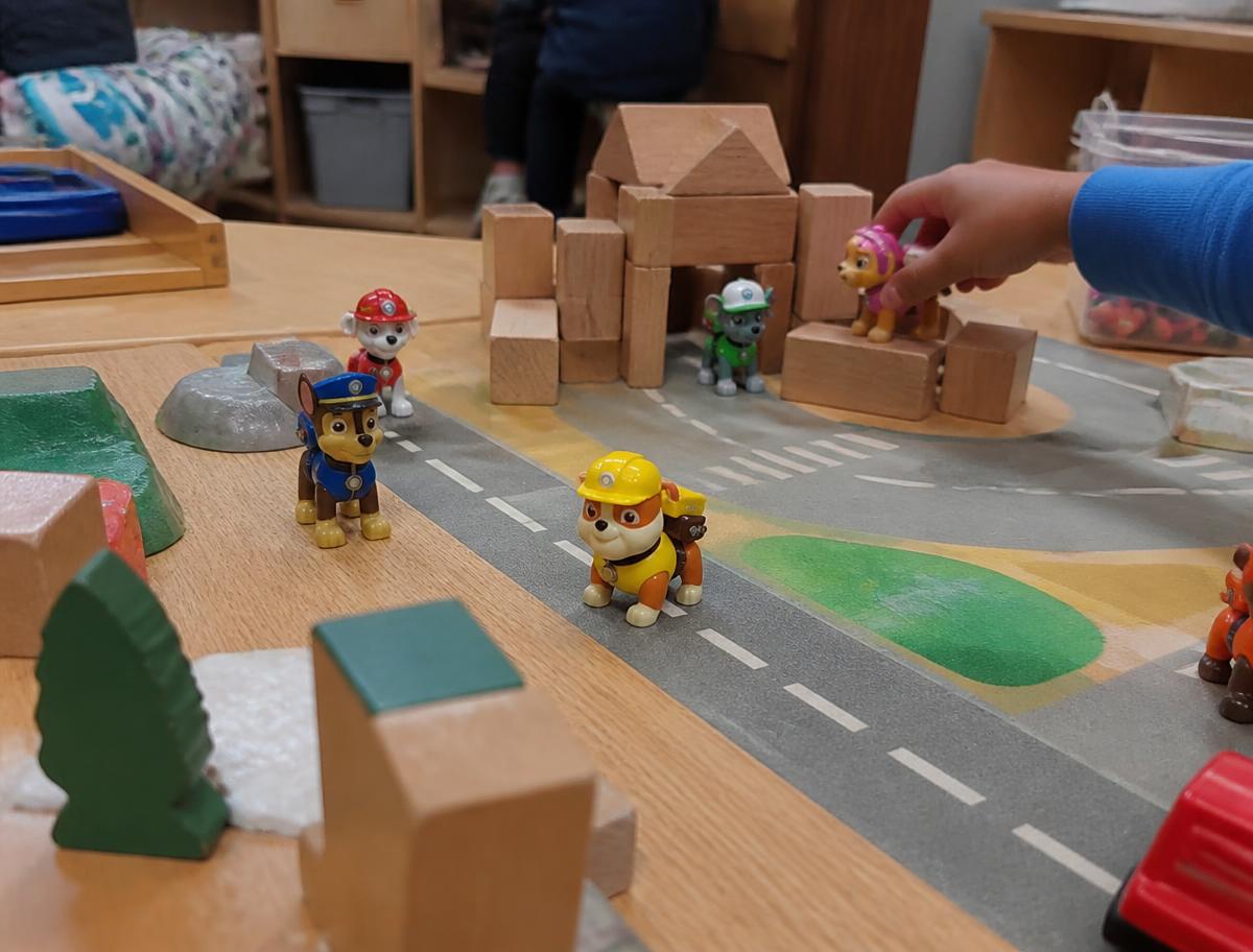 Paw Patrol figurines arranged in children's imaginative play with wooden blocks and a road mat on a timber classroom table